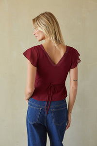A woman shown from the back wearing a burgundy short-sleeve wrap-style top with a lace-trimmed V-neck and tie waist, paired with blue jeans, against a plain beige wall.