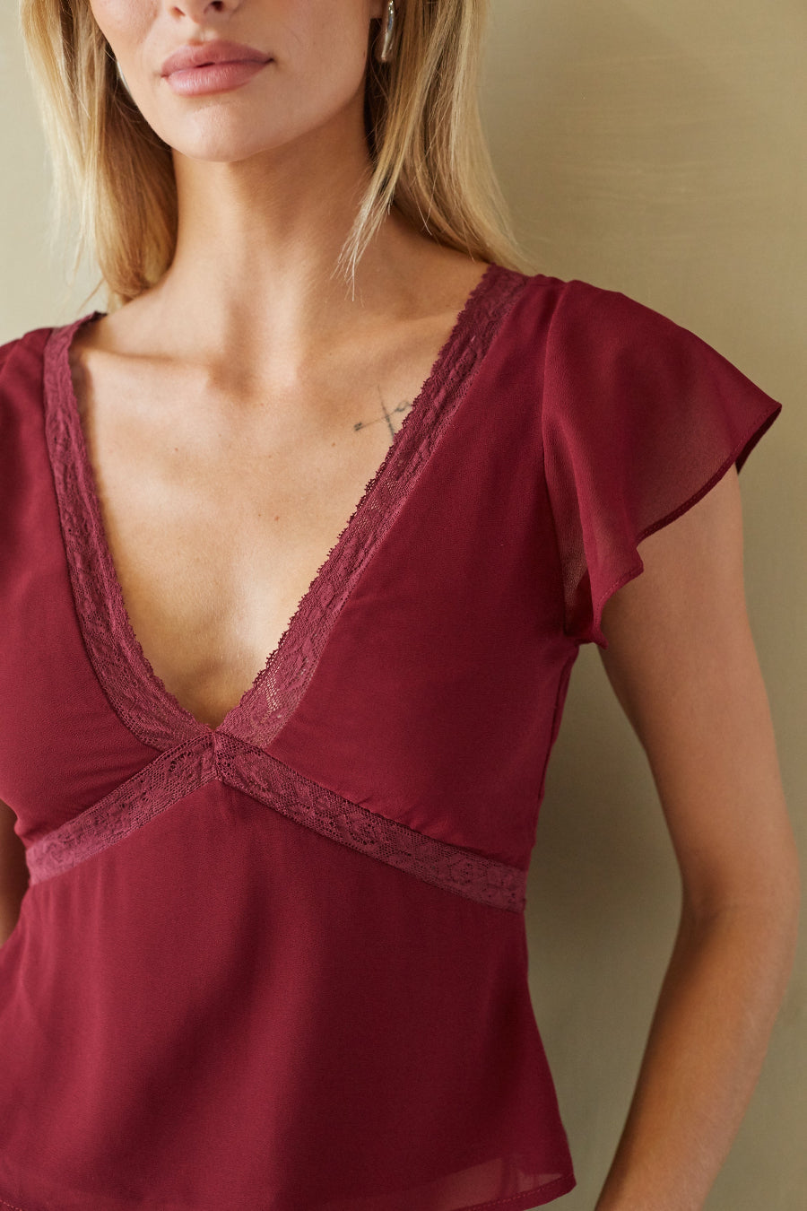 A close-up of a woman wearing a burgundy lace-trimmed camisole top with flutter sleeves, set against a neutral beige background.