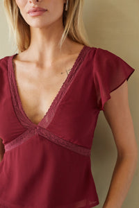 A close-up of a woman wearing a burgundy lace-trimmed camisole top with flutter sleeves, set against a neutral beige background.