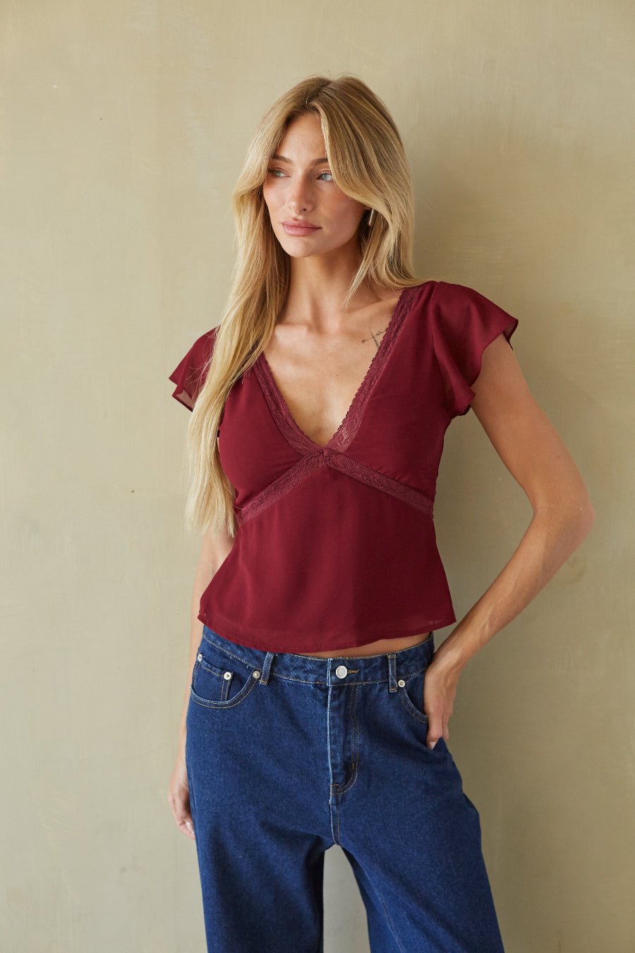 A woman modeling a burgundy short-sleeve top with a deep V-neck and lace trim, paired with blue jeans, standing against a textured beige wall.