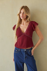 A woman modeling a burgundy short-sleeve top with a deep V-neck and lace trim, paired with blue jeans, standing against a textured beige wall.