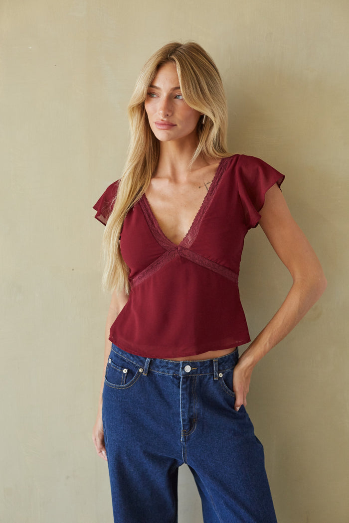 A woman modeling a burgundy short-sleeve top with a deep V-neck and lace trim, paired with blue jeans, standing against a textured beige wall.