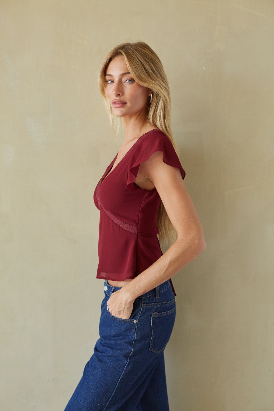 A woman modeling a short-sleeve wrap-style top in deep burgundy, paired with blue jeans, standing against a textured beige wall.