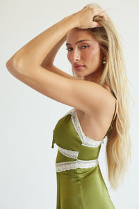A blonde woman posing in a satin olive green slip dress with white lace trim, set against a plain light gray background.
