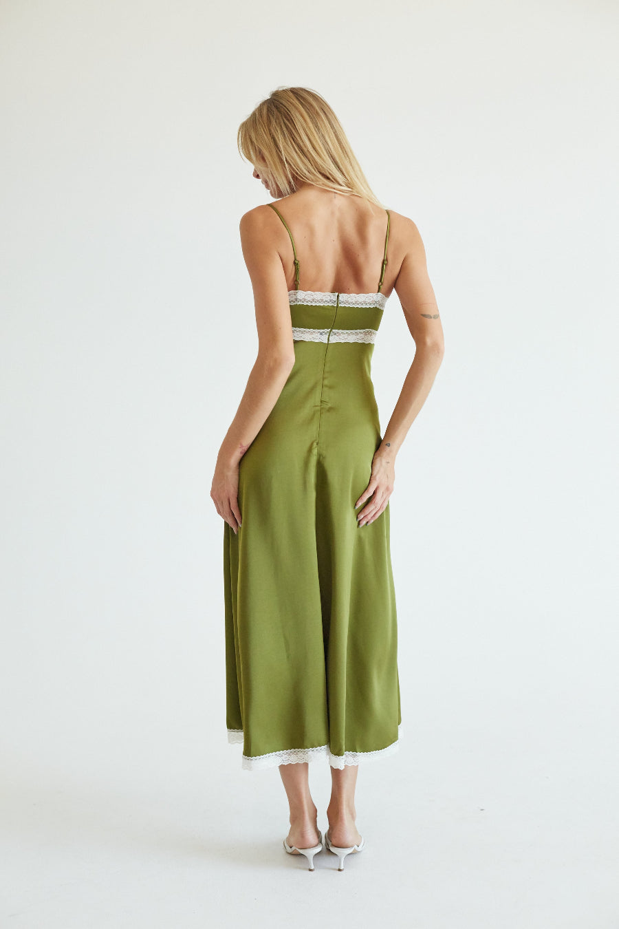A woman viewed from behind wearing an olive green midi slip dress with thin straps, a lace-trimmed back, and white heels, set against a plain white studio background.