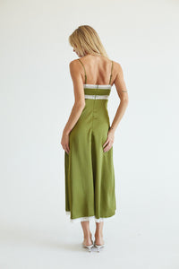 A woman viewed from behind wearing an olive green midi slip dress with thin straps, a lace-trimmed back, and white heels, set against a plain white studio background.