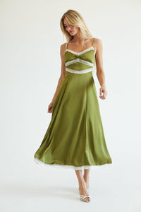 A woman modeling a flowing olive green midi dress with white lace trim at the bust, waist, and hem, photographed against a plain white studio background.