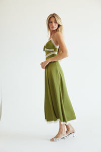 A woman modeling a sleeveless olive green maxi dress with white lace trim and white shoulder straps, paired with clear high-heeled sandals, against a plain white studio background.