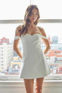 white-image | Woman wearing a white off-shoulder dress standing in front of a window with a cityscape view.