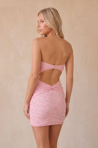 A woman modeling a pink strapless mini dress from the back, featuring a cutout across the back and a textured, embellished fabric, set against a neutral beige background.