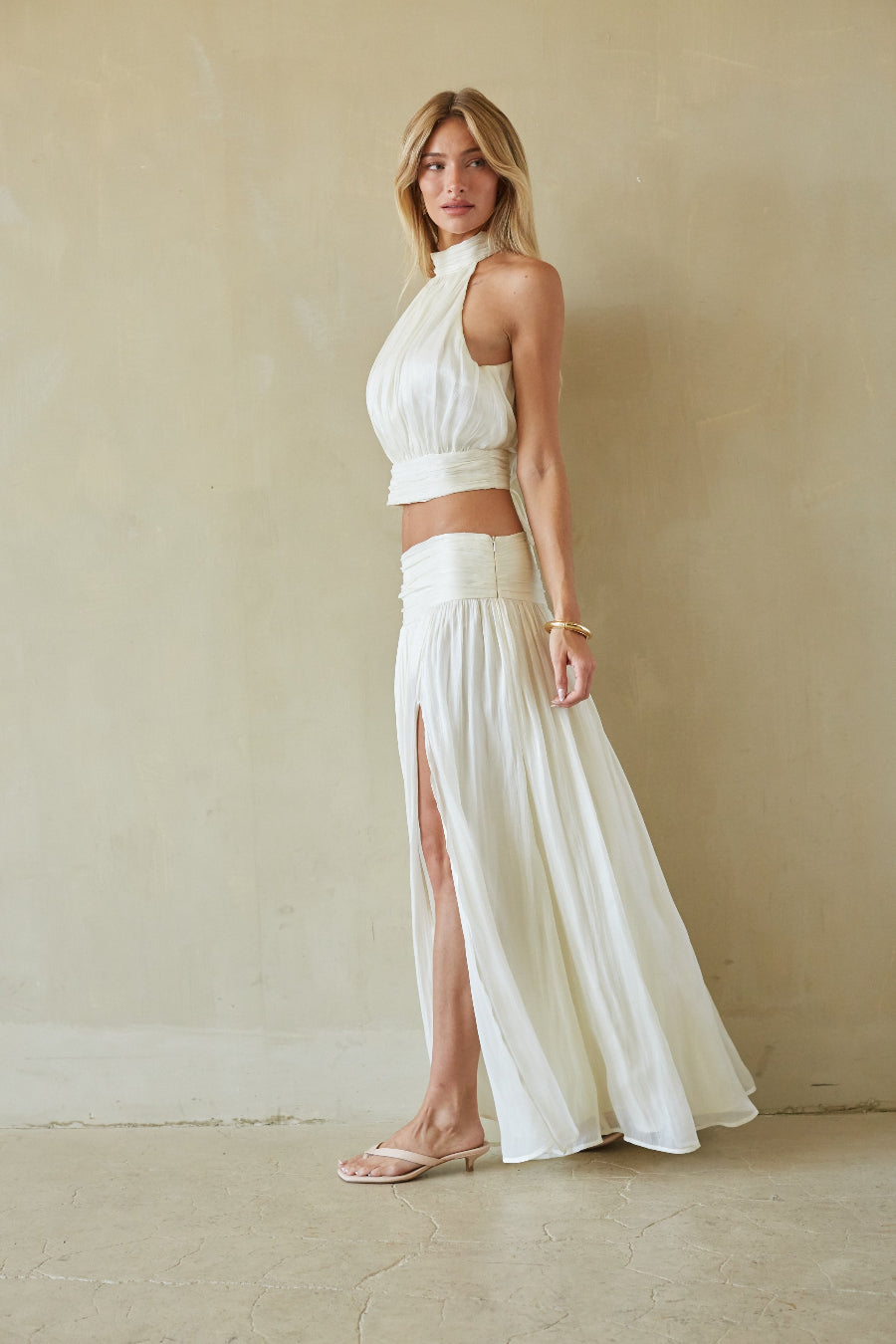 A woman modeling a two-piece white outfit consisting of a sleeveless halter crop top and a high-waisted maxi skirt with a thigh-high slit, standing against a textured beige plaster wall.