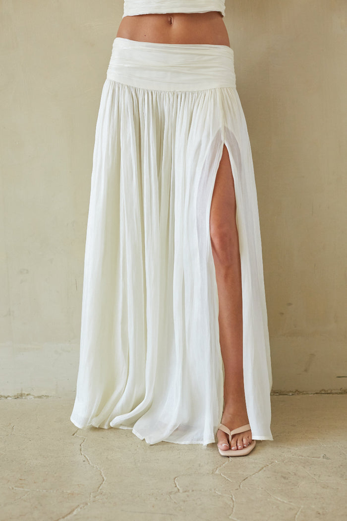 A person wearing a flowing white maxi skirt with a high side slit, paired with a white crop top and beige flat sandals, standing against a textured beige wall and floor.