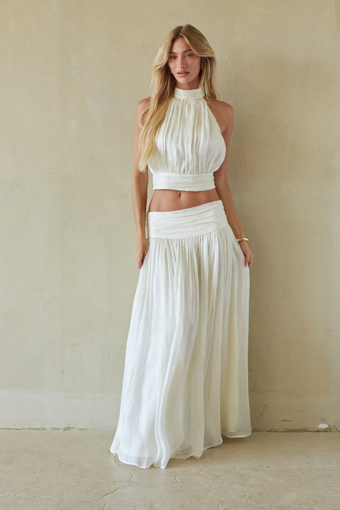 A woman modeling a two-piece white outfit consisting of a sleeveless halter crop top and a matching high-waisted pleated maxi skirt, standing against a textured beige wall.