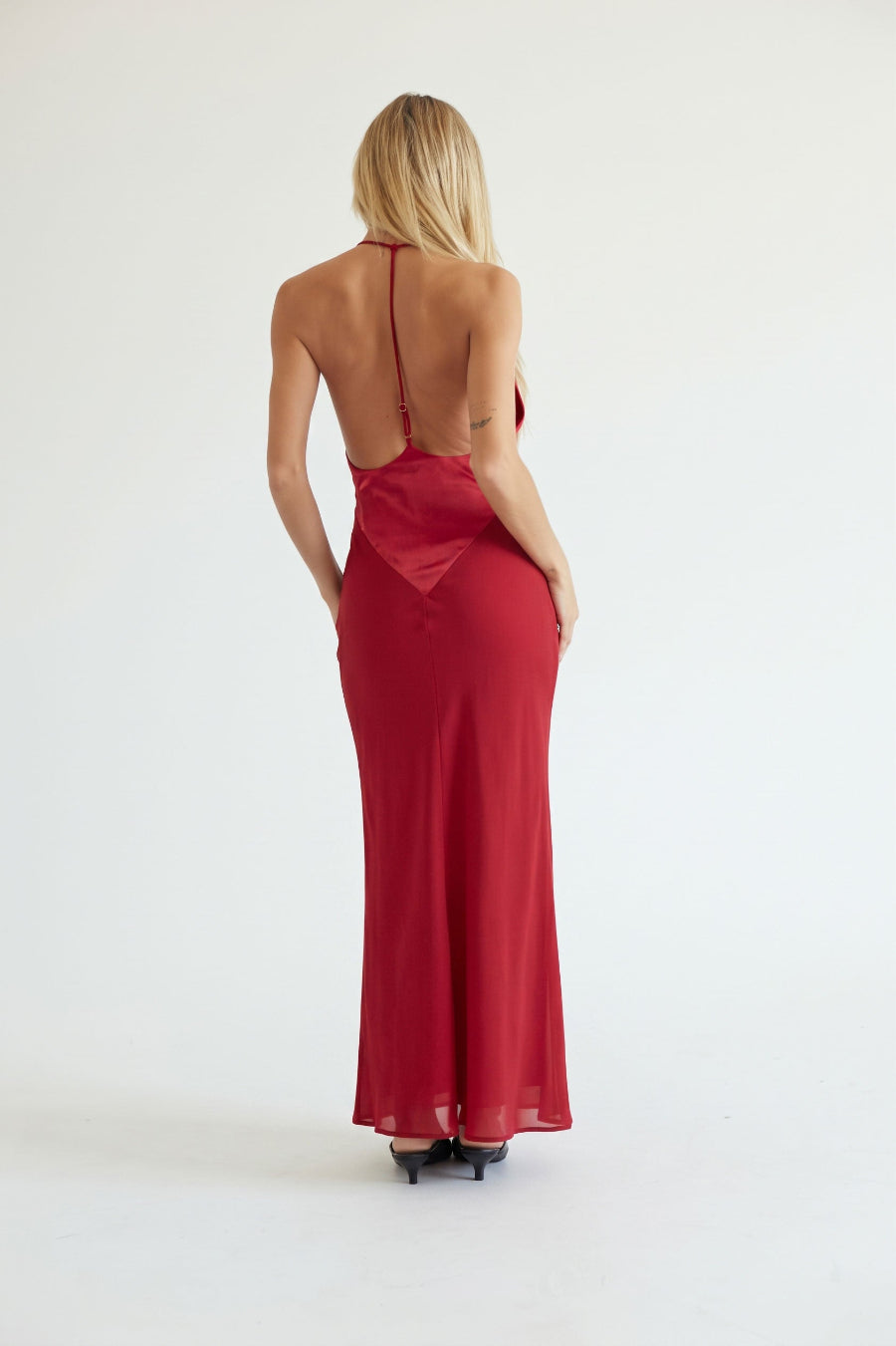 A woman viewed from behind wearing a long red satin slip dress with a low open back, photographed against a plain white studio background.