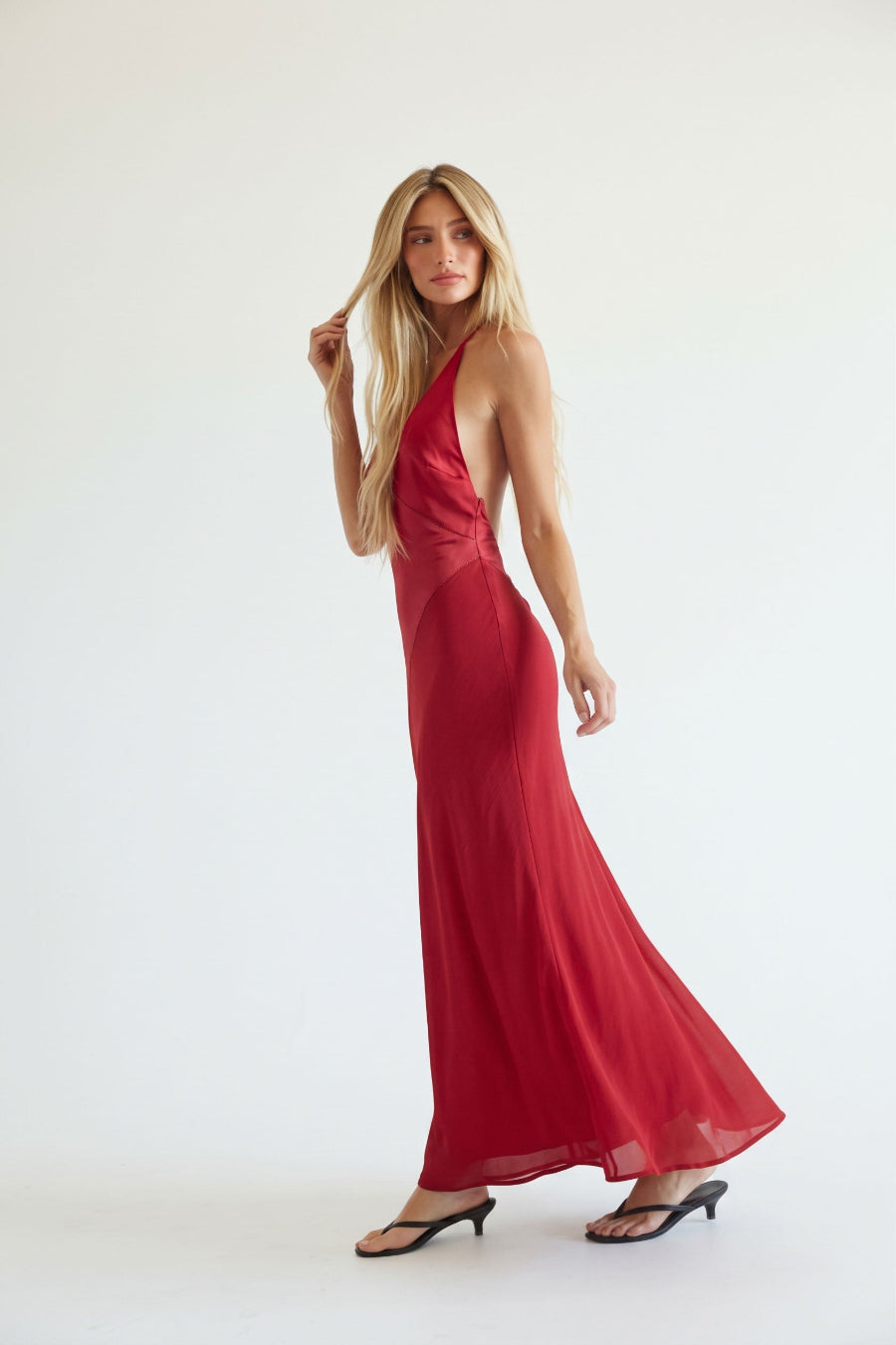 A woman modeling a long, flowing red halter-neck maxi dress with a draped, sheer fabric, paired with black heeled sandals against a plain white studio background.