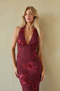 A blonde woman wearing a sleeveless deep burgundy halter-neck dress with 3D floral applique, standing against a textured beige wall.