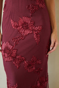 A close-up of a burgundy midi dress featuring 3D floral applique embroidery and sheer mesh side panels, worn by a model.