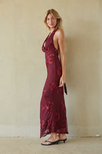 A woman modeling a sleeveless, deep burgundy maxi dress with a tonal floral lace overlay, standing against a textured beige plaster wall.