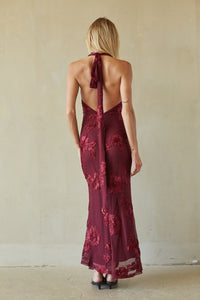 A woman viewed from behind wearing a sleeveless, open-back halter maxi dress in deep burgundy with tonal floral lace applique, standing against a textured beige wall.