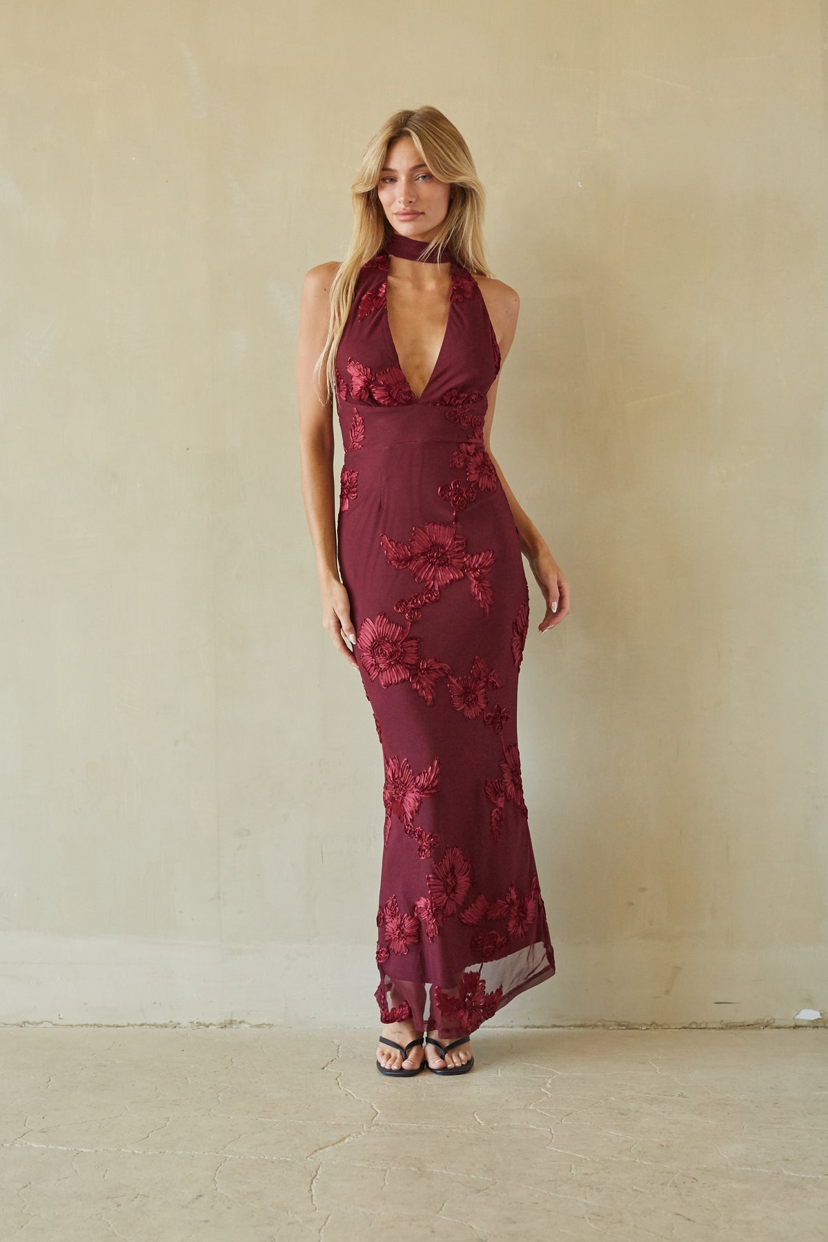 A woman modeling a sleeveless, deep V-neck, floor-length maroon lace dress with a choker neckline, standing against a textured beige plaster wall.