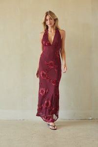 A woman modeling a sleeveless halter-neck maxi dress in deep burgundy with tonal floral lace applique, standing against a textured beige plaster wall.