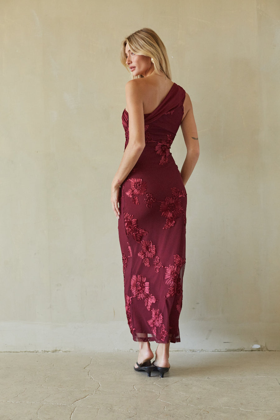 A woman wearing a sleeveless, one-shoulder burgundy maxi dress with floral lace applique, photographed from the back against a textured beige wall.