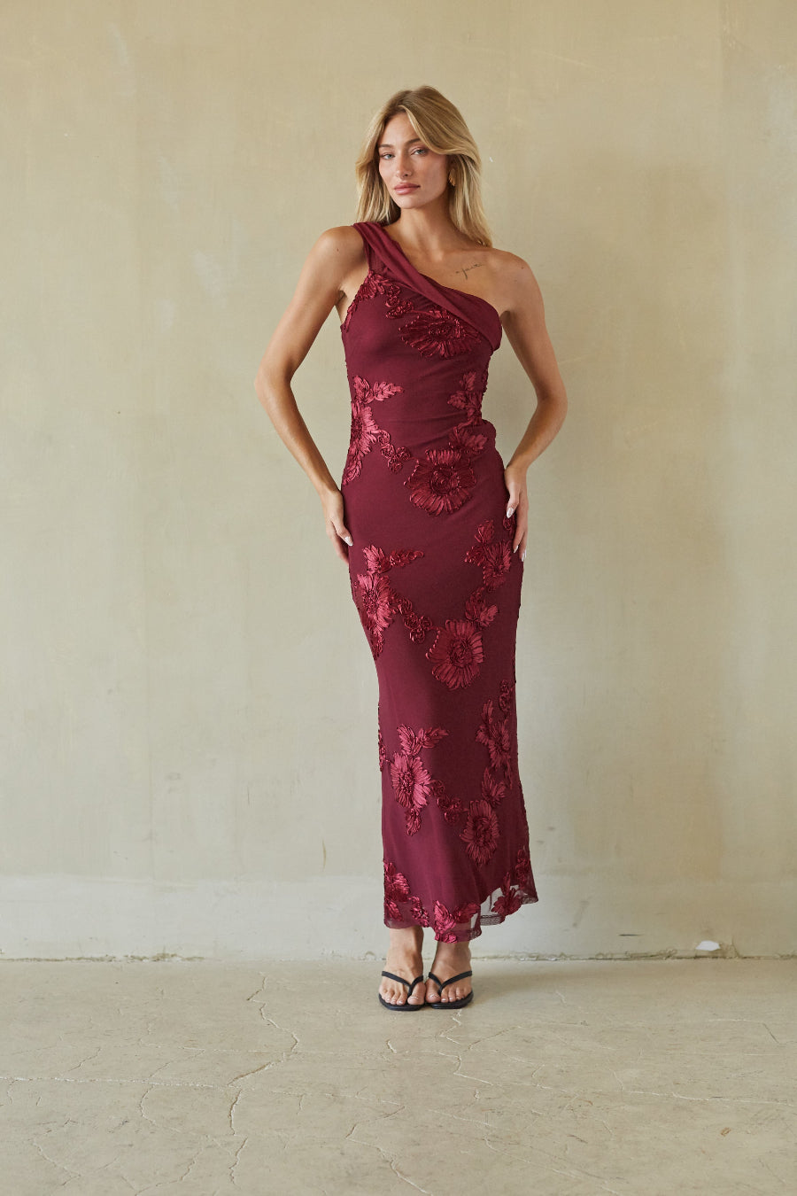 A woman modeling a one-shoulder, floor-length burgundy dress with a floral lace overlay, standing against a textured beige wall.