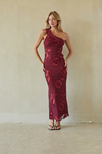 A woman modeling a one-shoulder, floor-length burgundy dress with a floral lace overlay, standing against a textured beige wall.