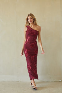 A woman modeling a one-shoulder midi dress in deep burgundy with a tonal floral lace overlay, standing against a textured beige wall.