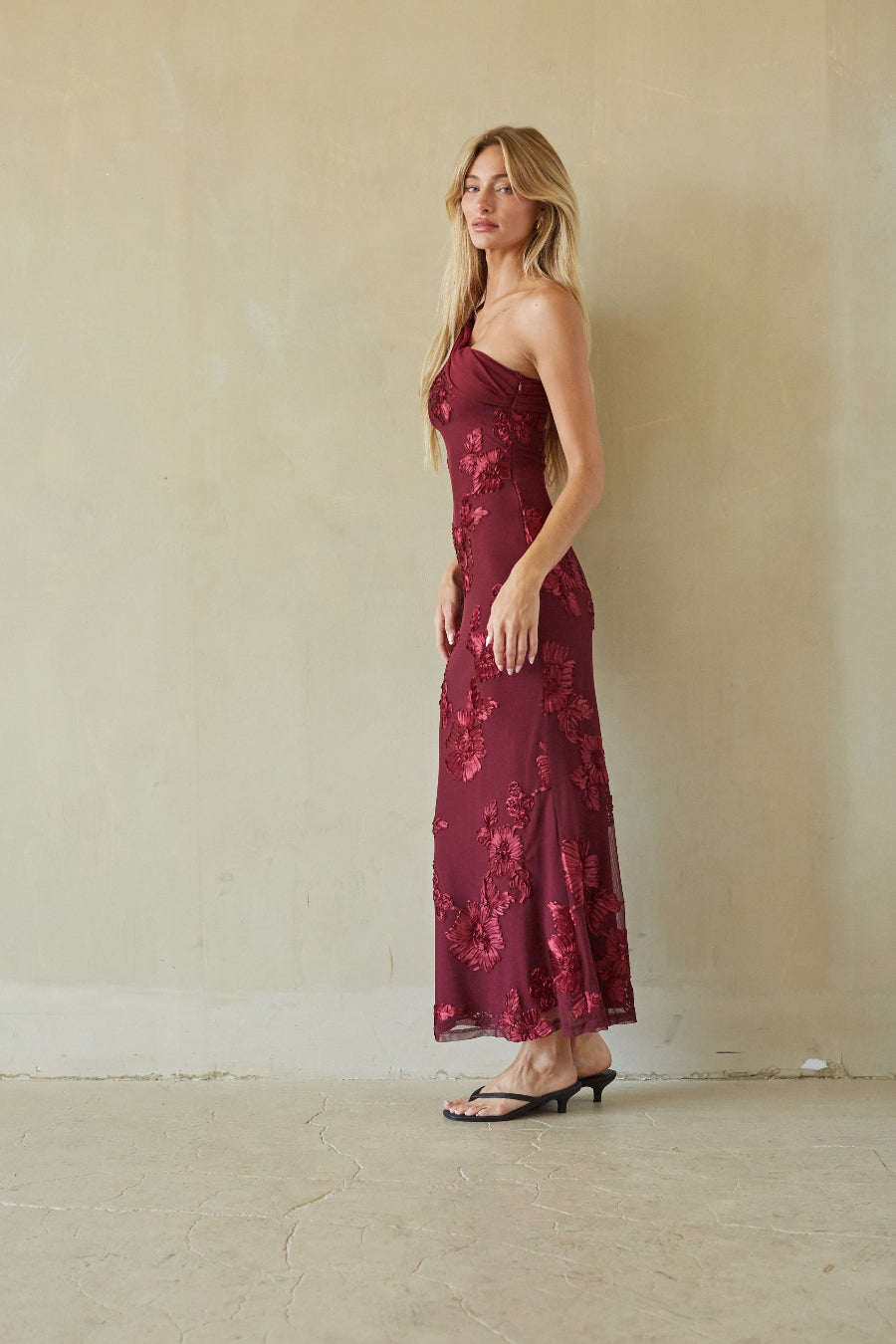 A woman modeling a strapless, floor-length burgundy maxi dress with a tonal floral pattern, standing against a textured beige plaster wall.