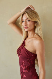 A blonde woman posing in a sleeveless, one-shoulder burgundy dress with floral embroidery, set against a neutral beige background.