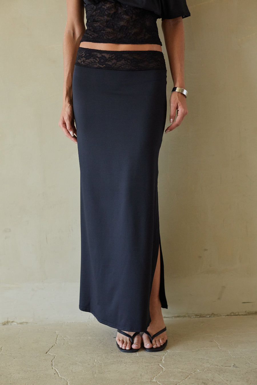 A woman wearing a long black satin maxi skirt with a side slit and a lace waistband, paired with black sandals, standing against a textured beige wall.