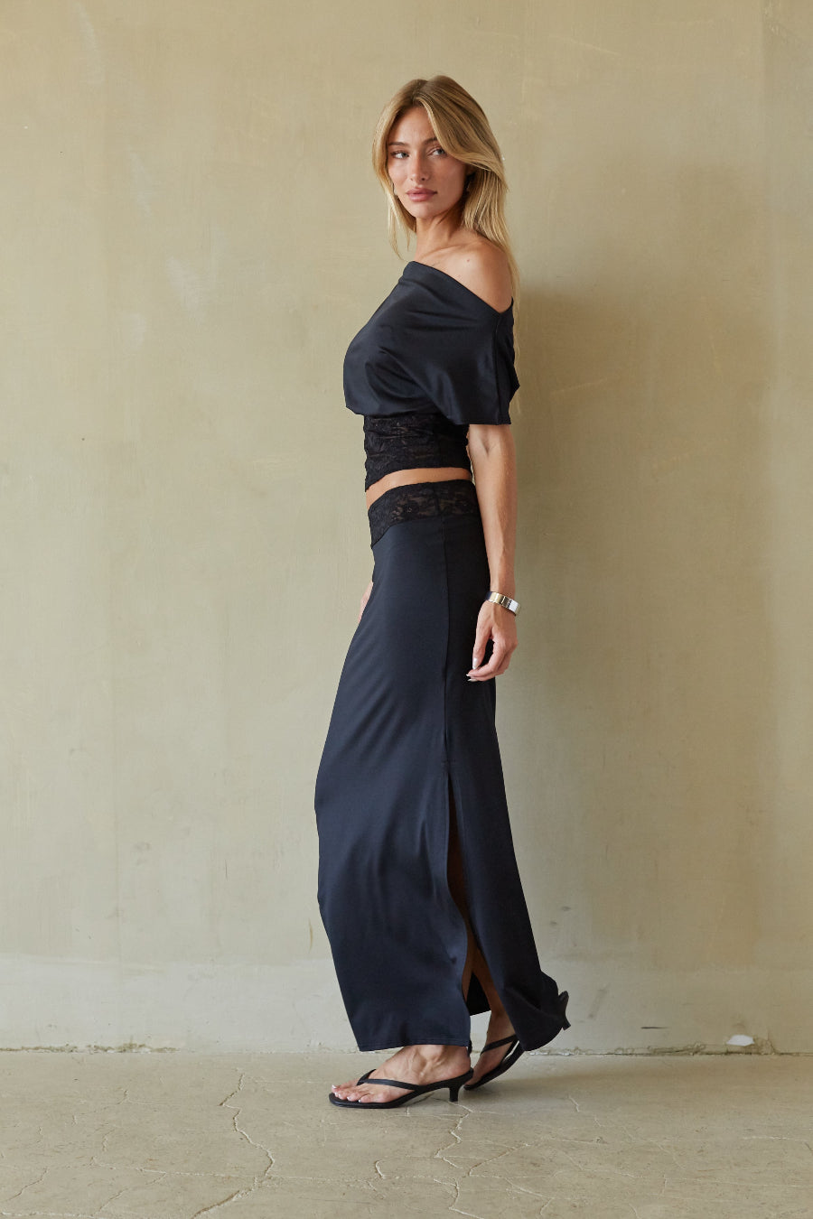 A woman models a two-piece navy blue outfit consisting of an off-the-shoulder crop top with black lace trim and matching high-waisted wide-leg skirt with a front slit, standing against a textured beige wall.