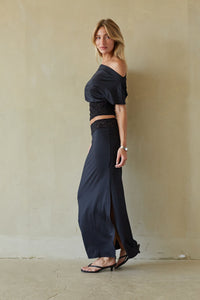 A woman models a two-piece navy blue outfit consisting of an off-the-shoulder crop top with black lace trim and matching high-waisted wide-leg skirt with a front slit, standing against a textured beige wall.