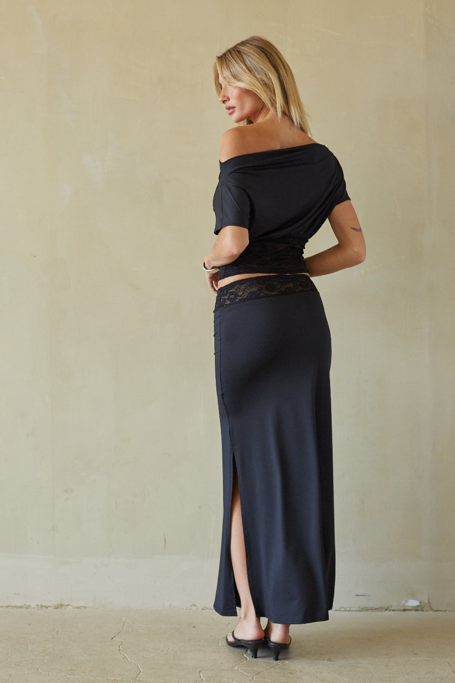 A woman modeling a dark navy off-the-shoulder maxi dress with a high side slit and a black lace waistband, standing against a neutral beige wall.