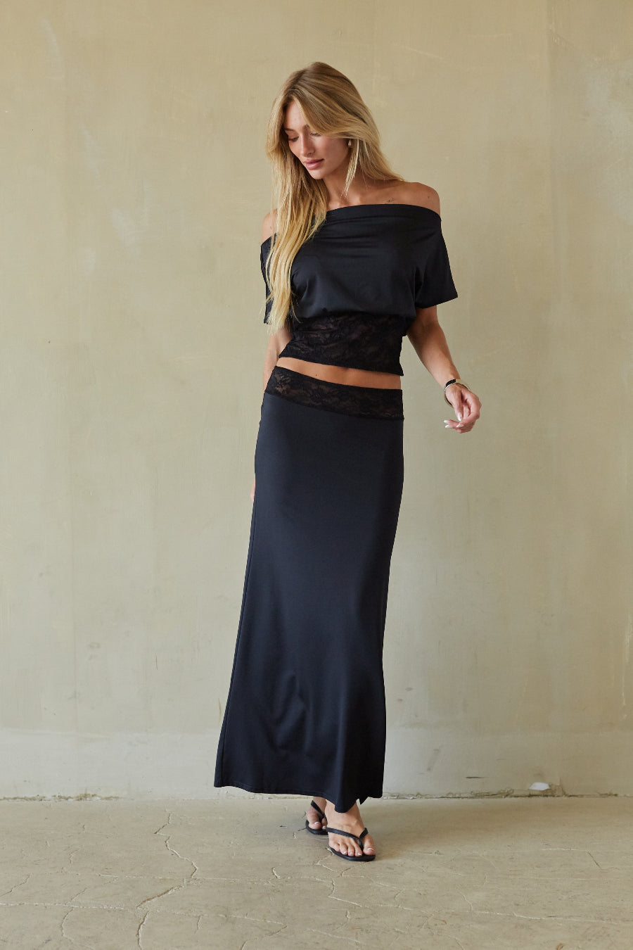 A woman modeling a black off-the-shoulder crop top with a black lace panel and a matching long black skirt, standing against a textured beige wall.