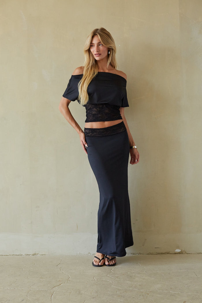 A woman modeling a two-piece navy blue outfit consisting of an off-the-shoulder crop top with a black lace panel and a matching high-waisted maxi skirt, standing against a textured beige wall.