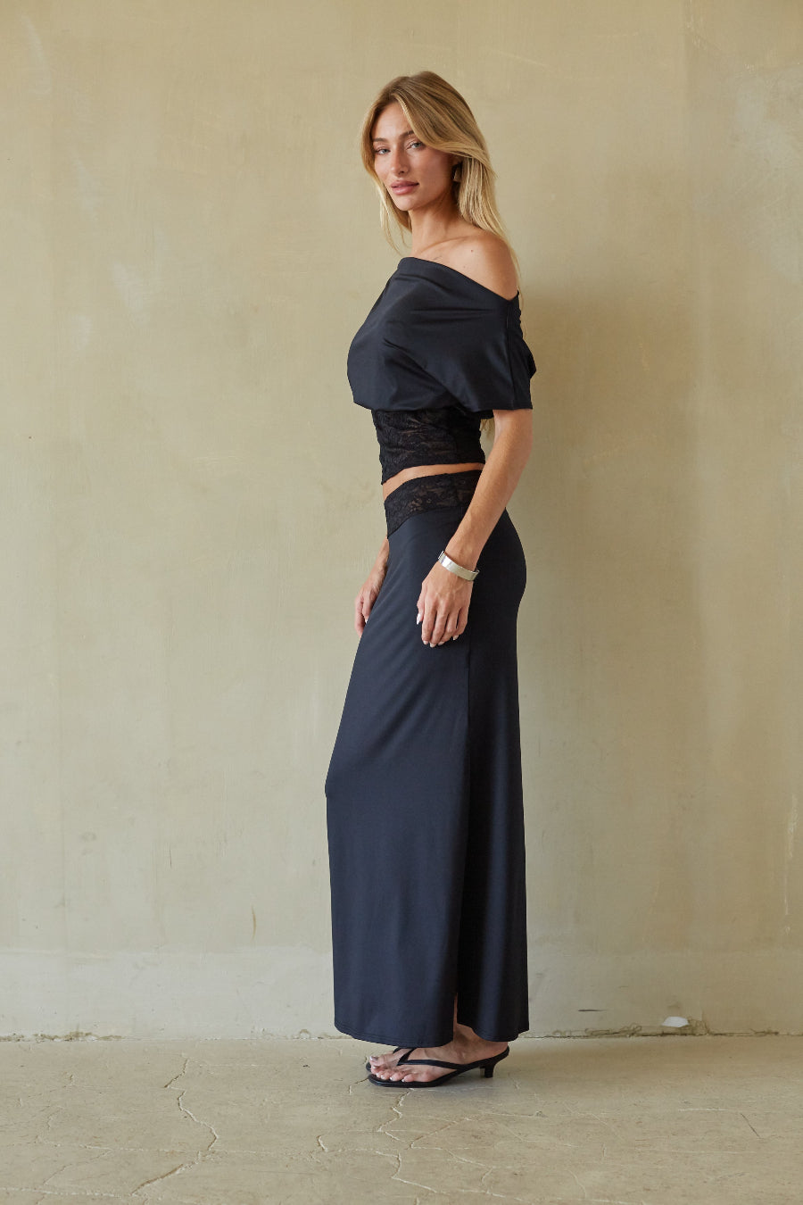 A woman modeling a dark navy off-the-shoulder maxi dress with a black lace-trimmed crop top, standing against a textured beige plaster wall.