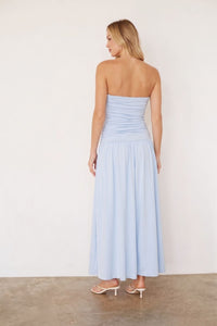 A woman wearing a light blue strapless maxi dress, shown from the back against a plain off-white wall and concrete floor.