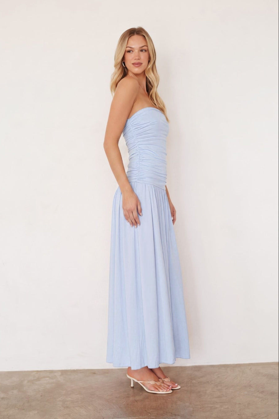 A woman modeling a strapless light blue maxi dress with a ruched bodice and flowing skirt, standing against a plain off-white wall with a concrete floor.