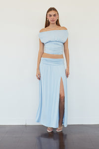 A woman modeling a light blue two-piece outfit consisting of an off-the-shoulder crop top and a matching high-slit maxi skirt, standing against a plain white wall.