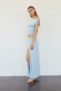 A woman models a light blue two-piece outfit consisting of a ruched crop top and a matching high-slit maxi skirt, standing against a plain white wall.