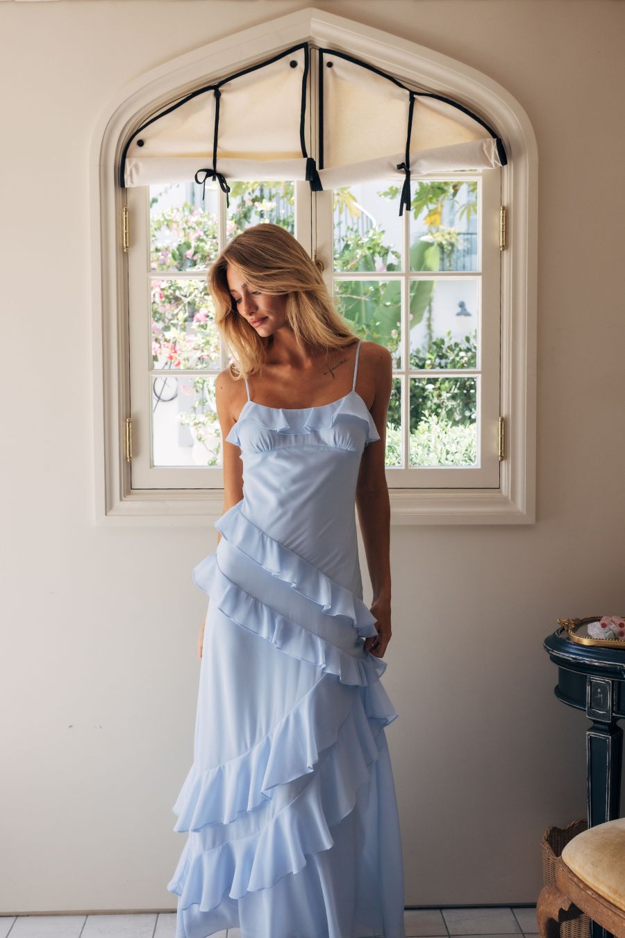 Woman in a light blue ruffled dress standing in a room with a large window.