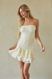 A blonde woman models a strapless, off-white sequined mini dress with a ruffled hem against a textured beige studio backdrop.