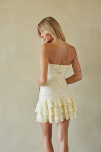 A woman wearing a strapless cream-colored mini dress with intricate beadwork and a tiered ruffle hem, photographed from behind against a textured beige wall.