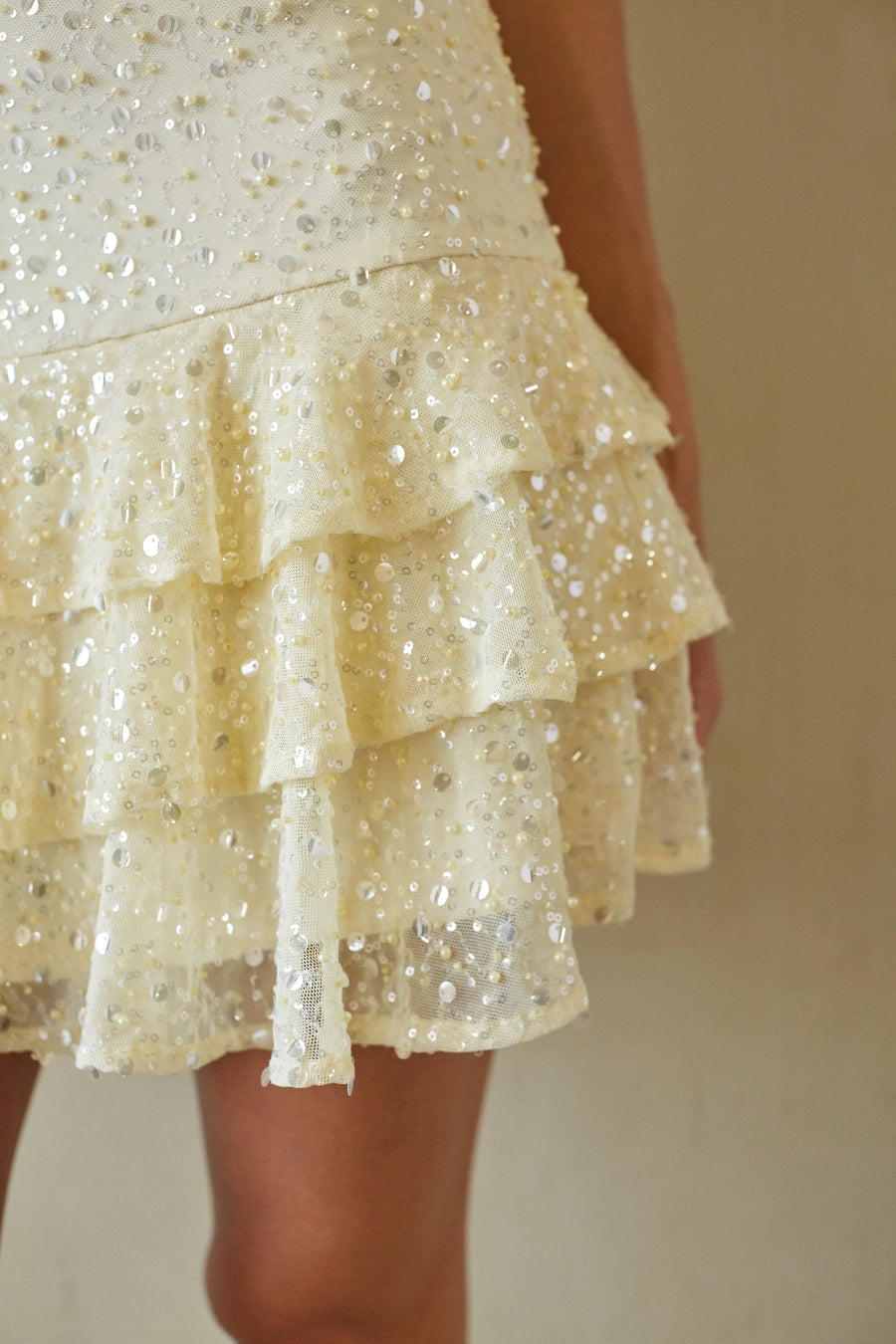 Close-up of a tiered ruffled skirt covered in shimmering silver and white sequins, worn by a model against a plain beige background.