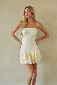 A blonde model wearing a strapless cream-colored mini dress with a textured bodice and a ruffled tiered skirt, posing against a neutral beige wall.