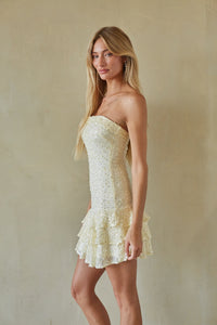 A blonde model wearing a strapless cream sequined mini dress with a ruffled tiered skirt, standing against a textured beige wall.