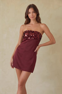 red-image | A woman modeling a strapless mini dress in deep burgundy with a sequined embellished bodice, set against a neutral beige textured background.