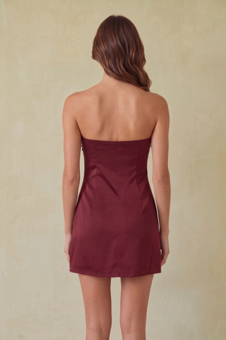 A woman viewed from the back wearing a strapless mini dress in a deep burgundy color, set against a textured beige background.